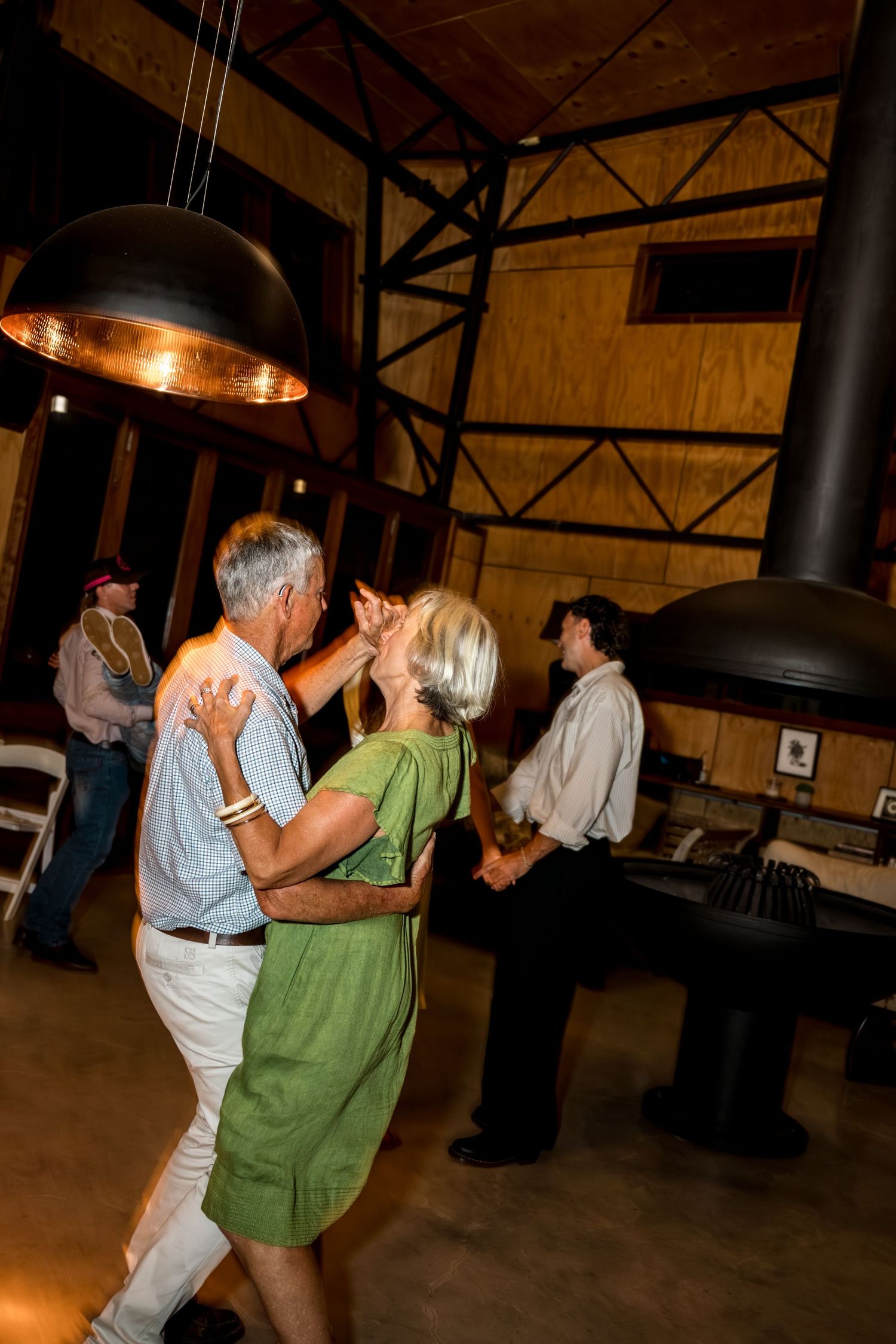 Older couple shares a romantic dance at a warmly lit rustic wedding reception.
