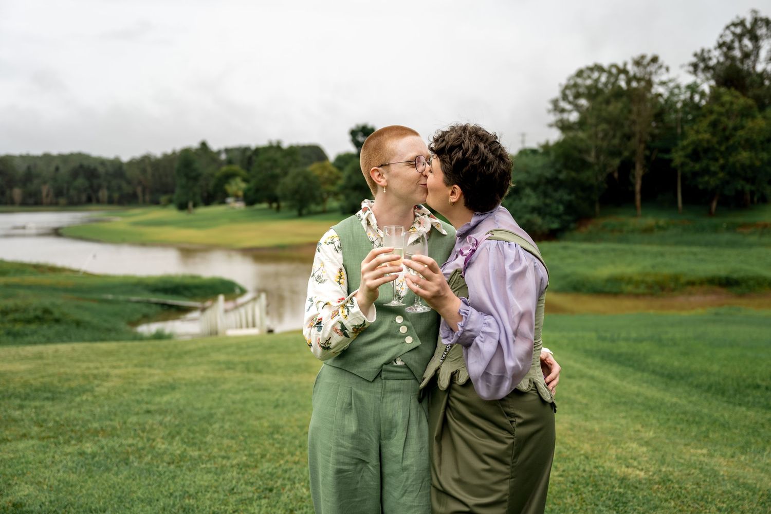 Couple in matching green outfits sharing a kiss while holding champagne glasses outdoors.