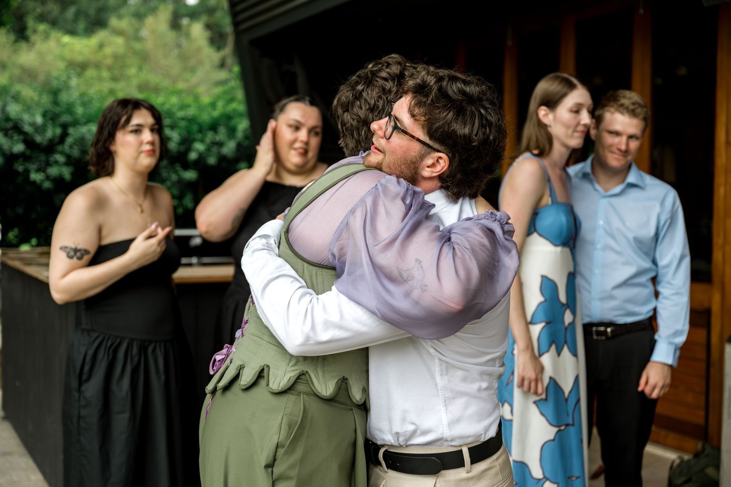 Two people share an emotional hug outside a wedding venue with guests watching.