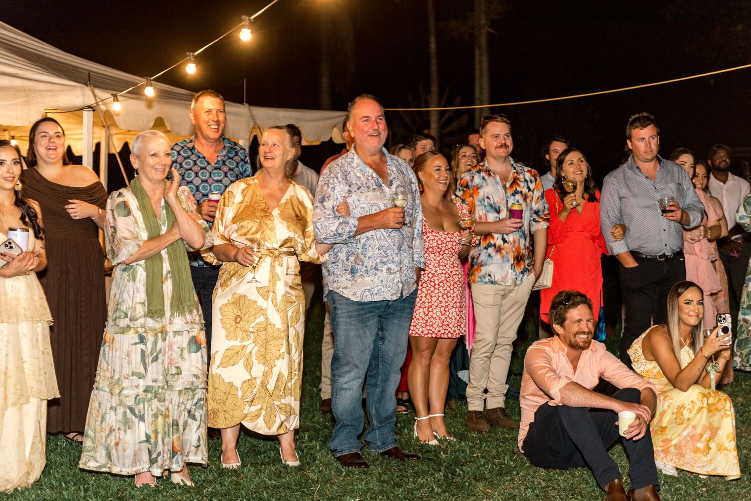 Large group of smiling wedding guests gathered outdoors under string lights near white marquee tent at evening reception.