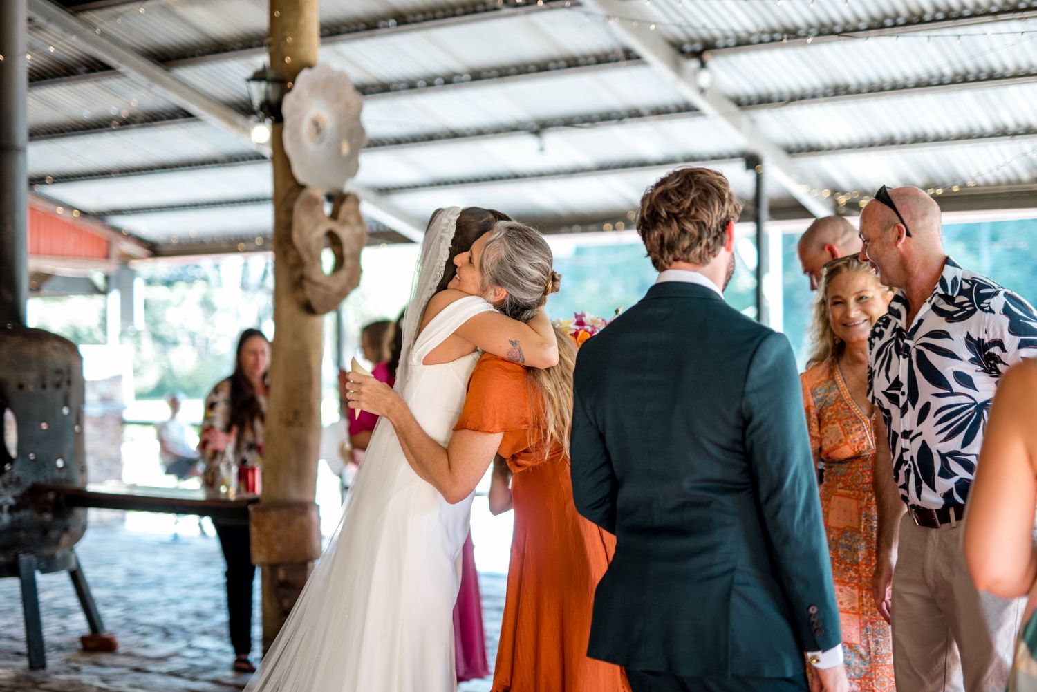 Bride in white gown hugs guest in orange dress as groom stands nearby at wedding reception.