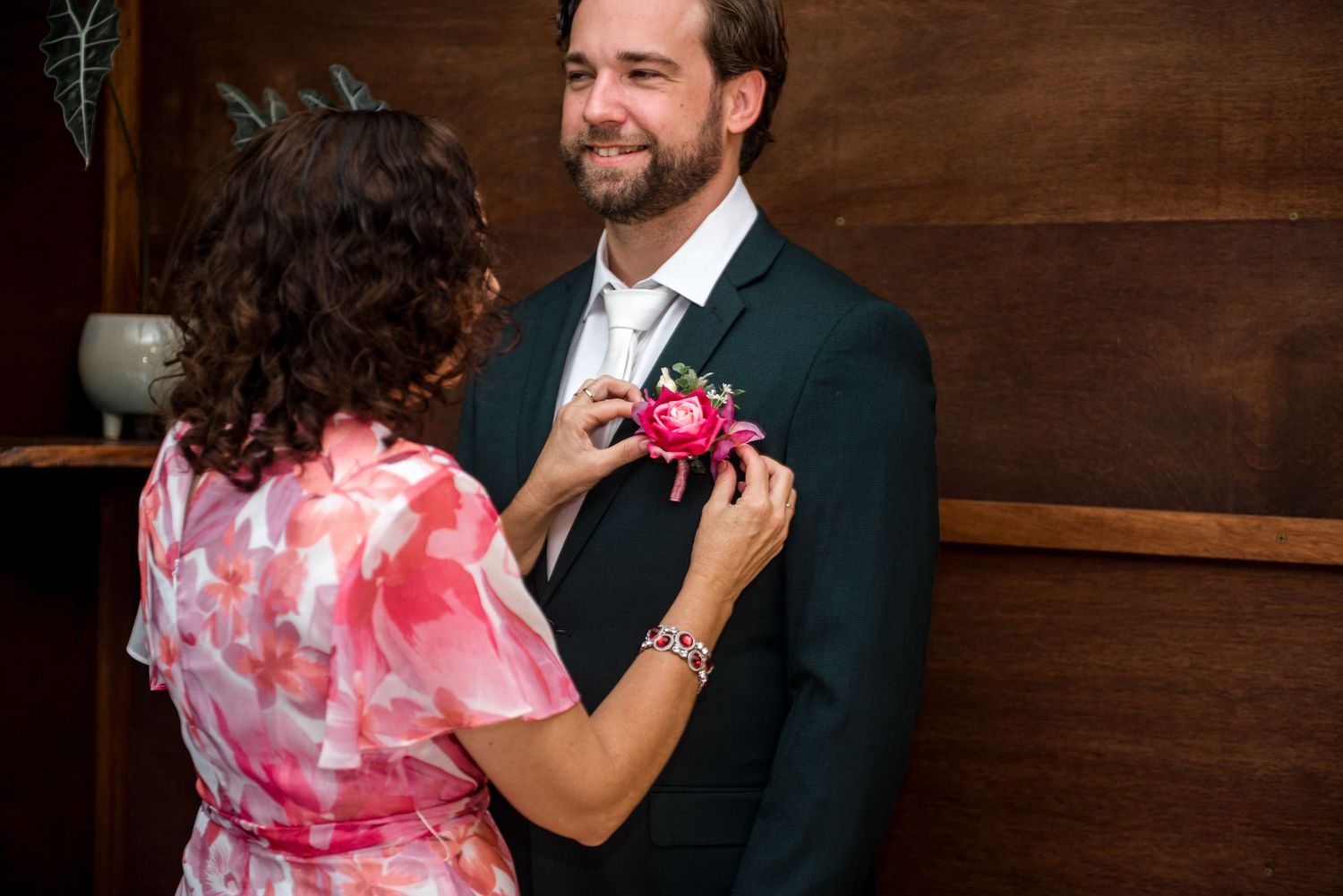 Mother pinning pink peony boutonniere on smiling groom in dark suit.