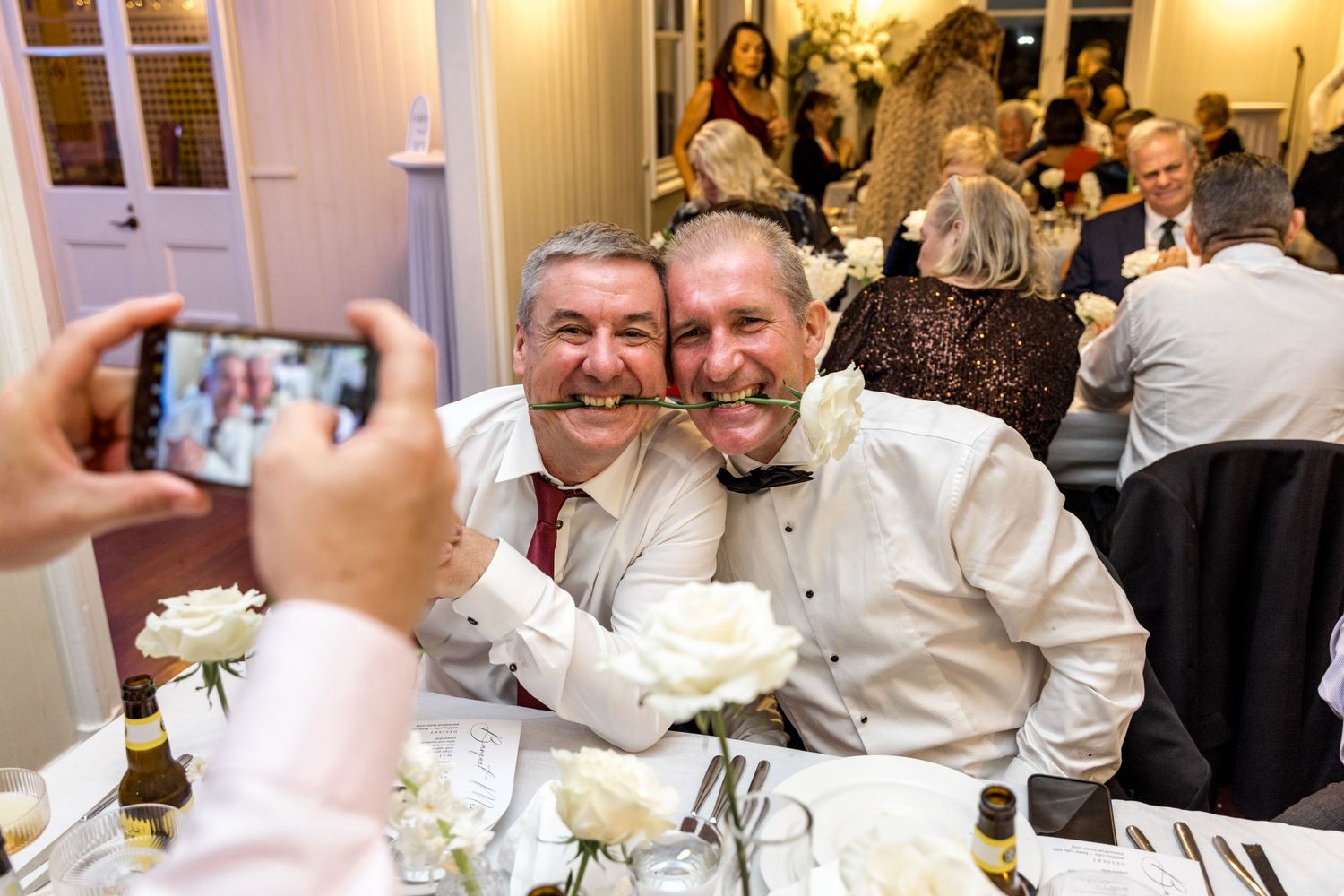 Two men posing playfully with a white rose at an elegant wedding reception dinner.