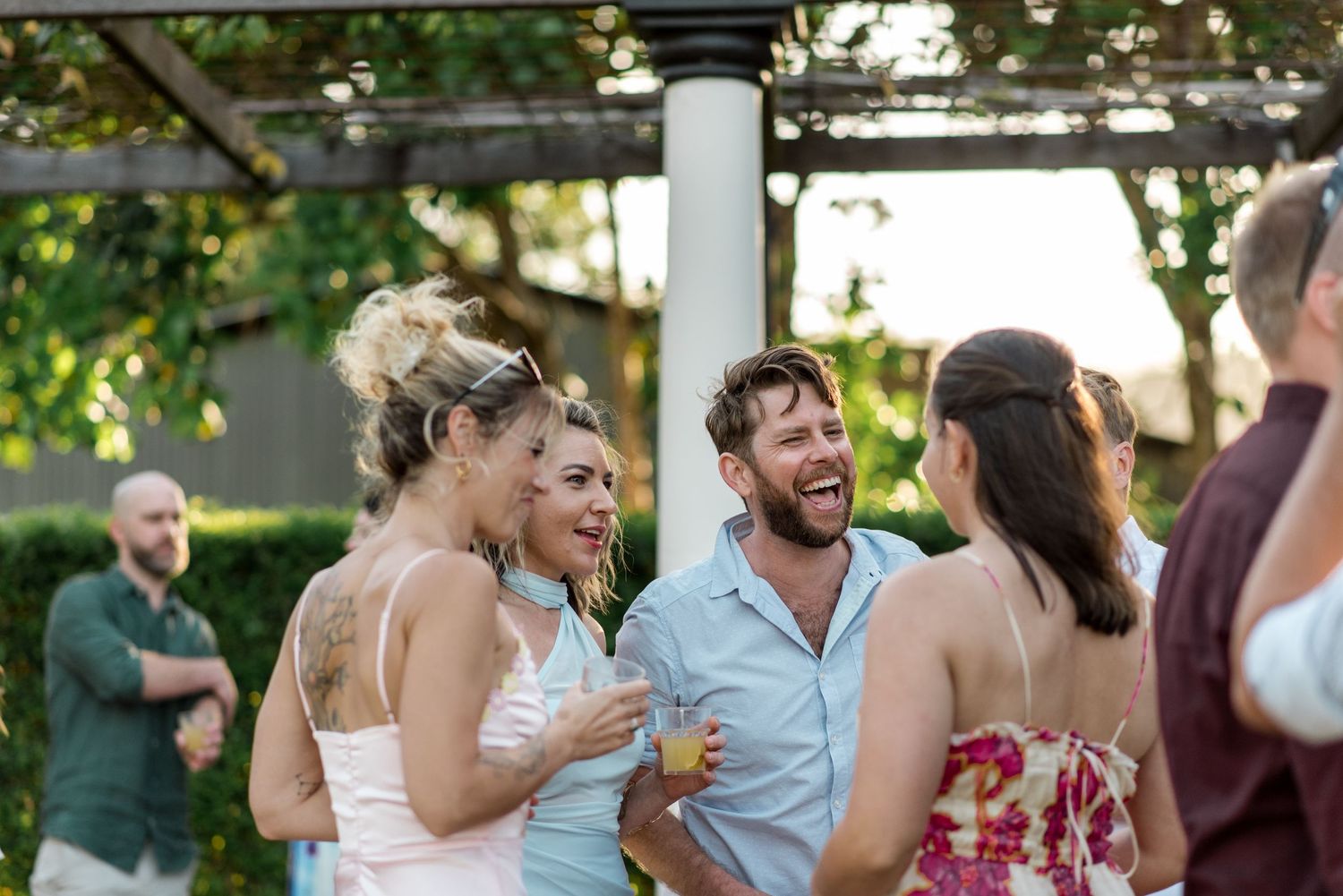 Wedding guests laughing and socializing outdoors under pergola during garden cocktail hour celebration.