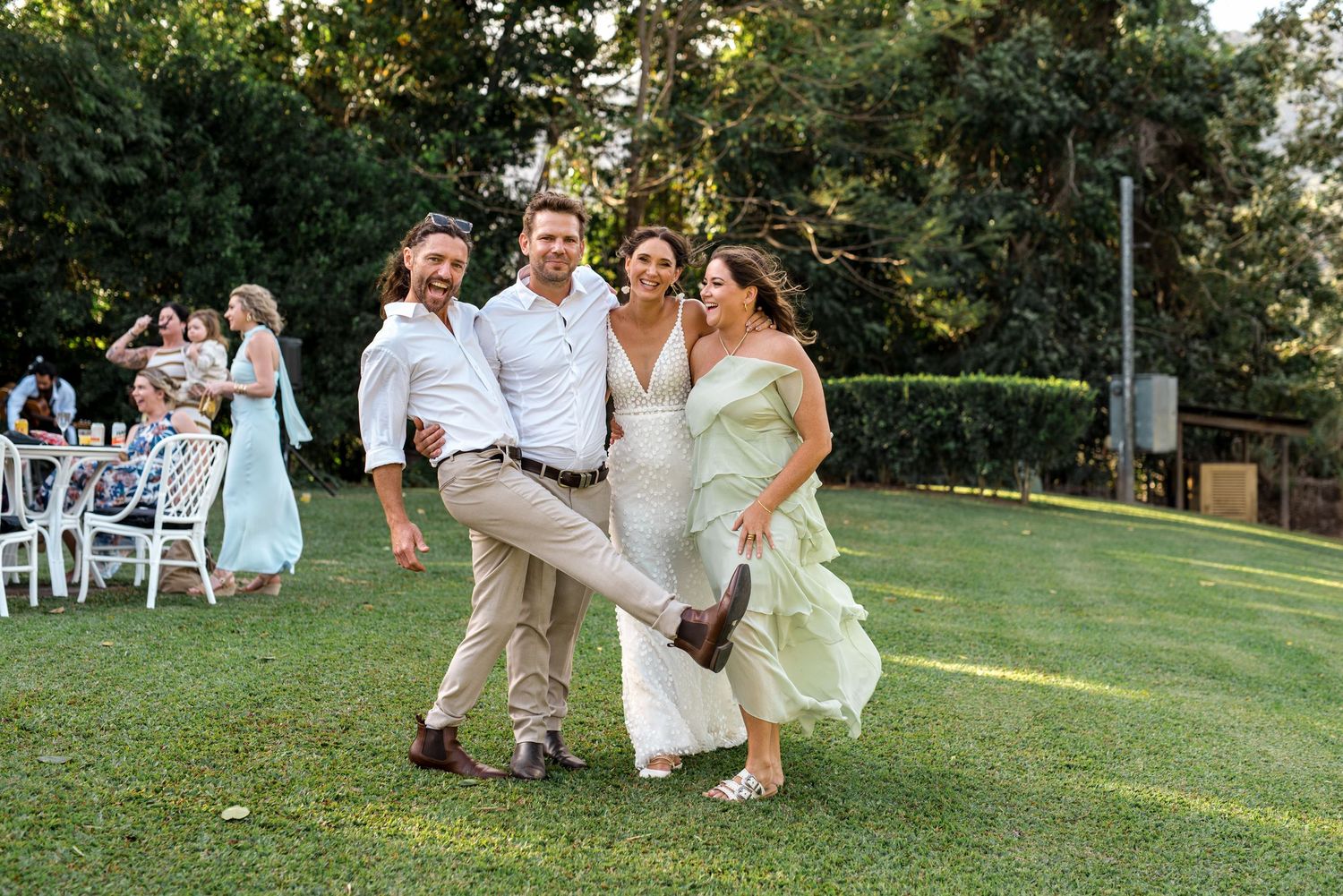 Joyful wedding guests laugh and pose together on green lawn at outdoor garden reception.