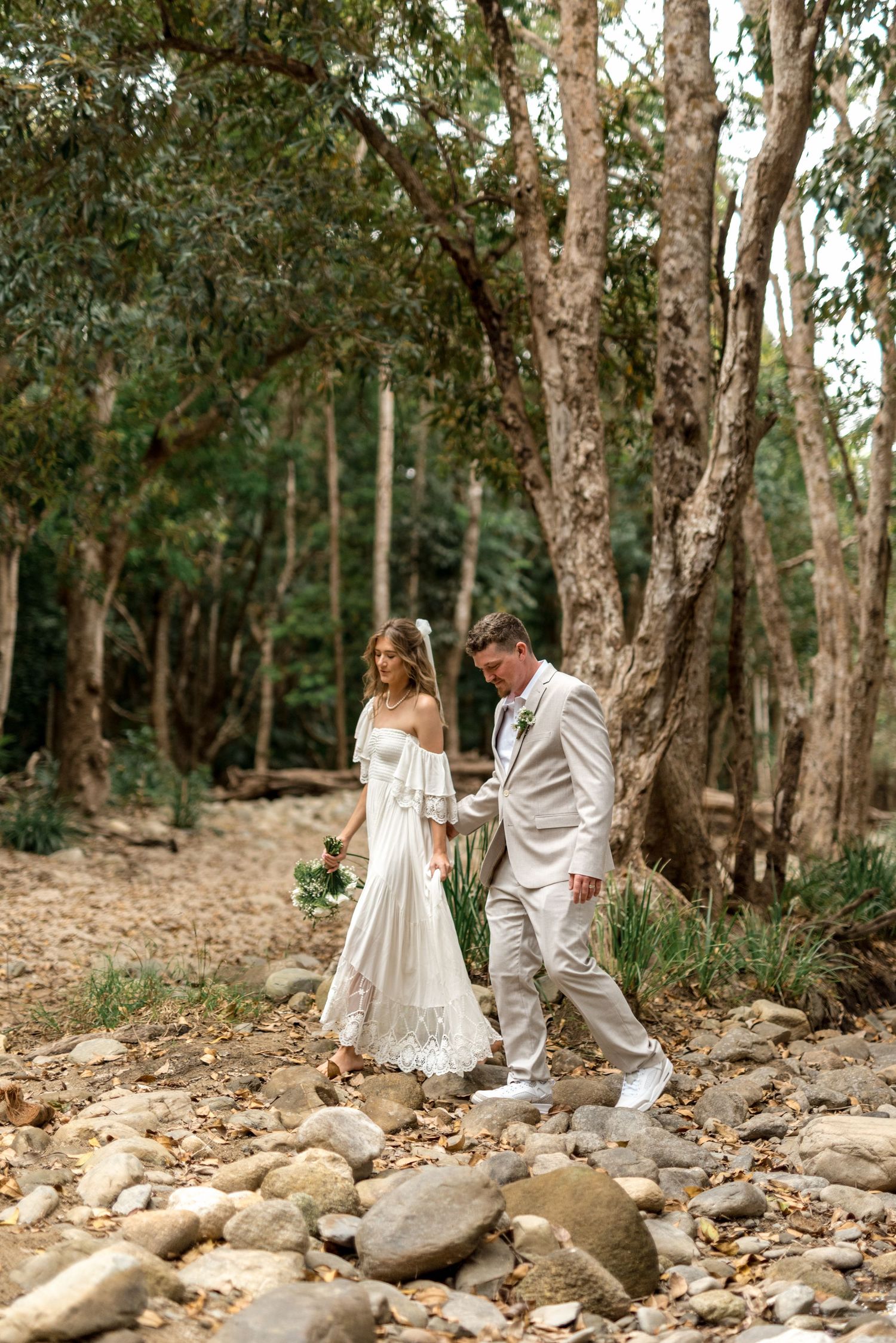 Bride in boho white dress and groom in tan suit walk hand in hand through a rocky forest.