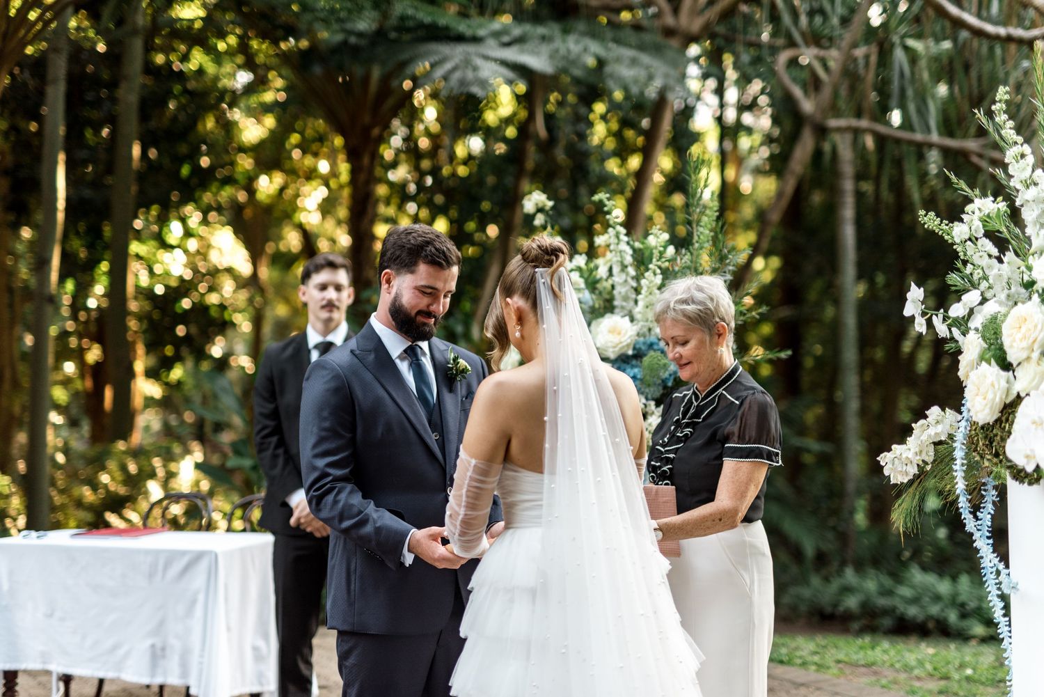 Bride and groom exchange vows during outdoor garden wedding ceremony with celebrant and lush greenery.