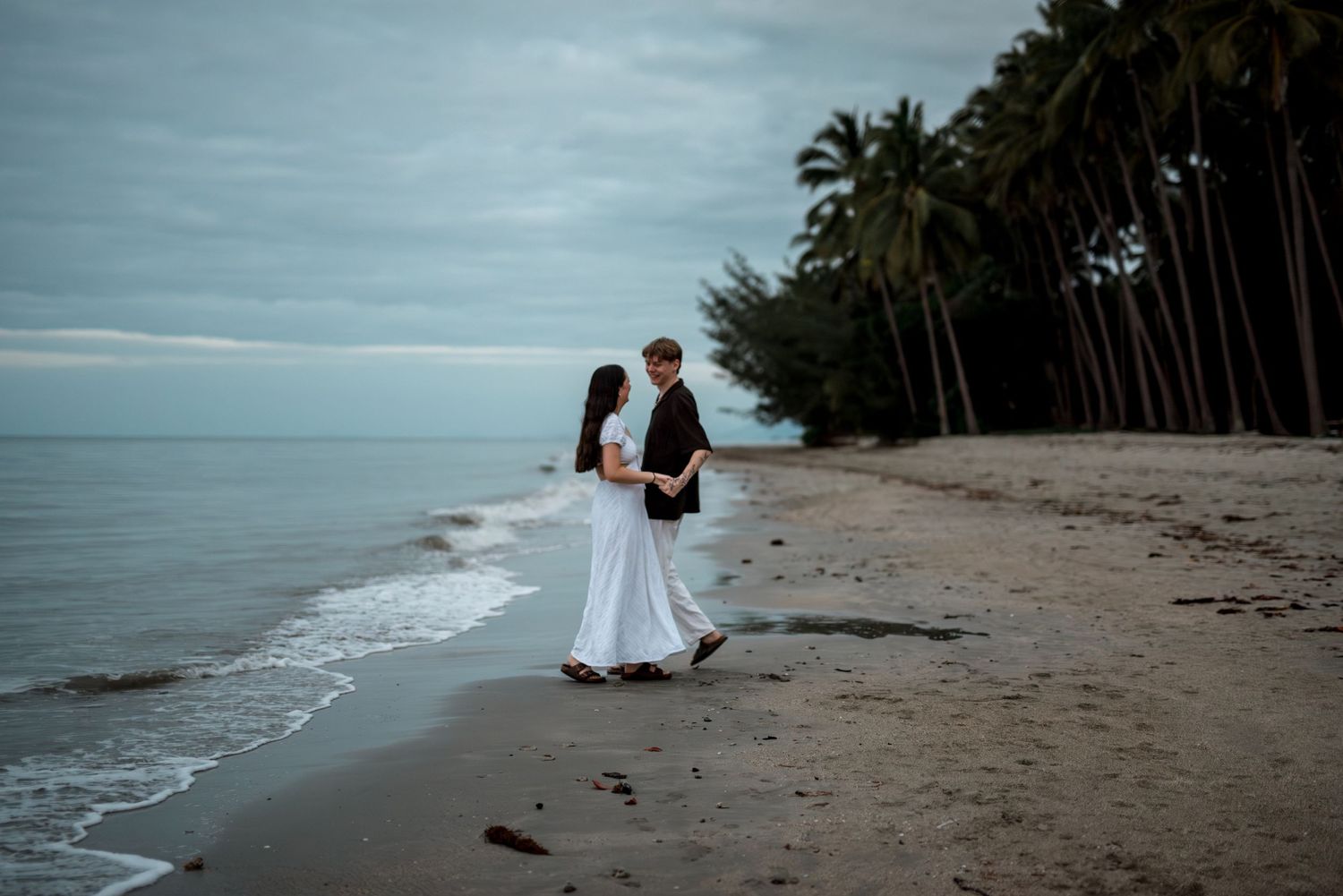 Couple walking hand in hand along a tropical beach with lush palm trees and cloudy skies.