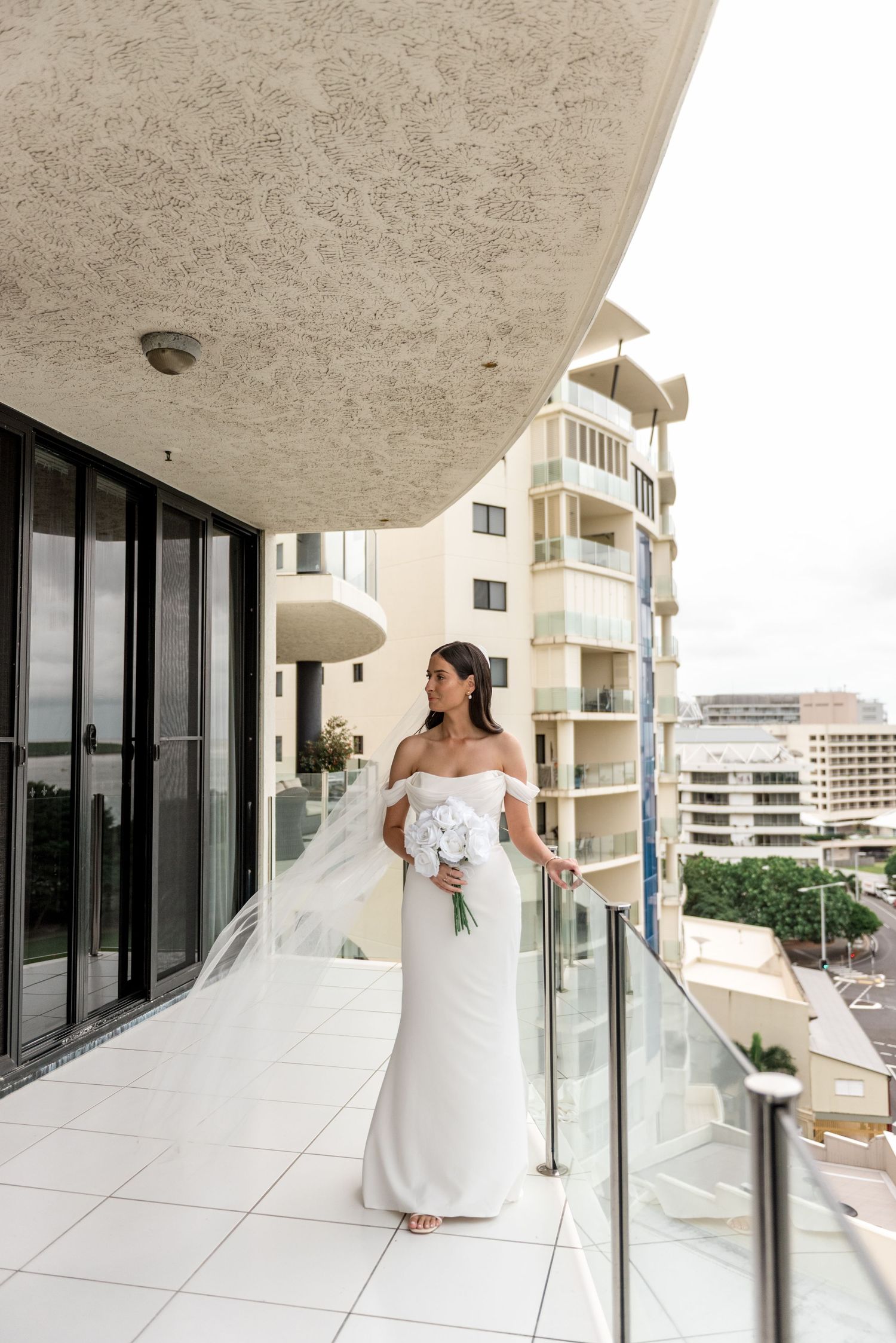 Bride in off-shoulder white gown holding white bouquet on high-rise hotel balcony.