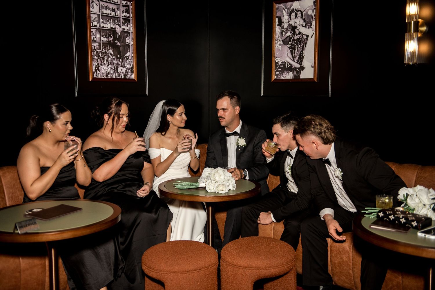 Bride and groom laugh with bridesmaids and groomsmen in a stylish dark lounge setting.