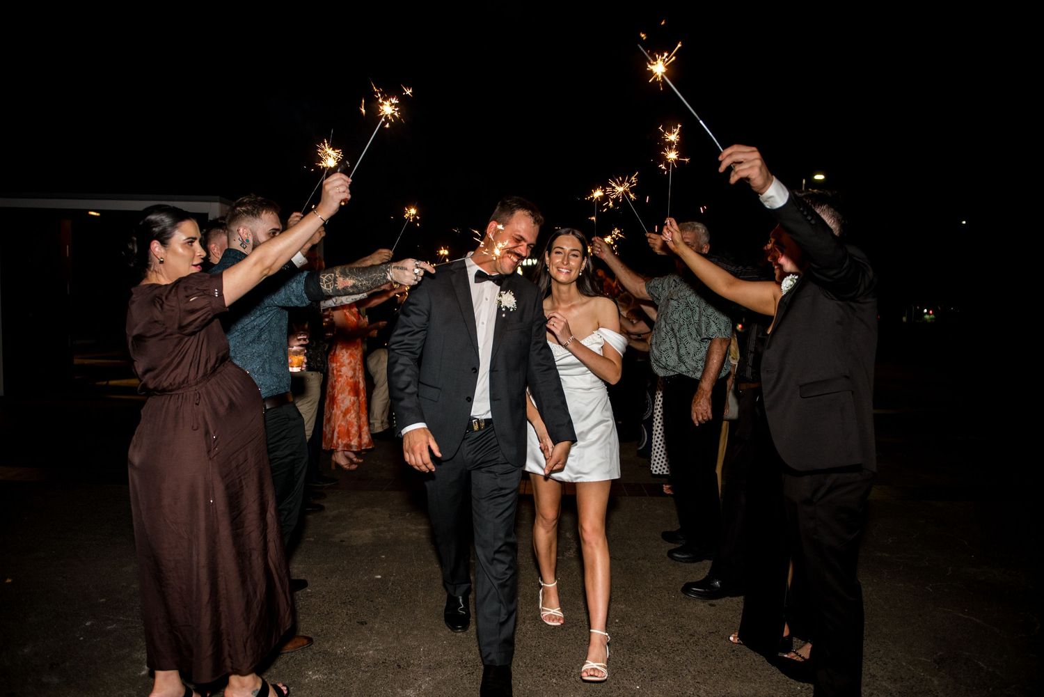 Bride and groom walk through a sparkler tunnel surrounded by cheering wedding guests at night.