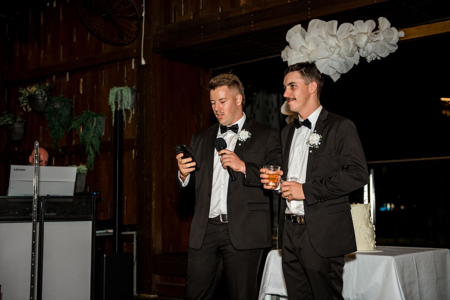 Two groomsmen in tuxedos deliver a wedding speech, one holding a phone and the other a drink, beside a white cake.