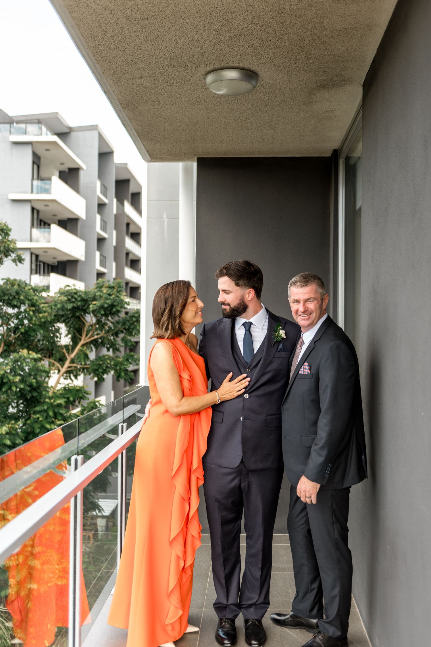 Groom in navy suit with boutonniere poses with smiling parents on a balcony overlooking apartment buildings.