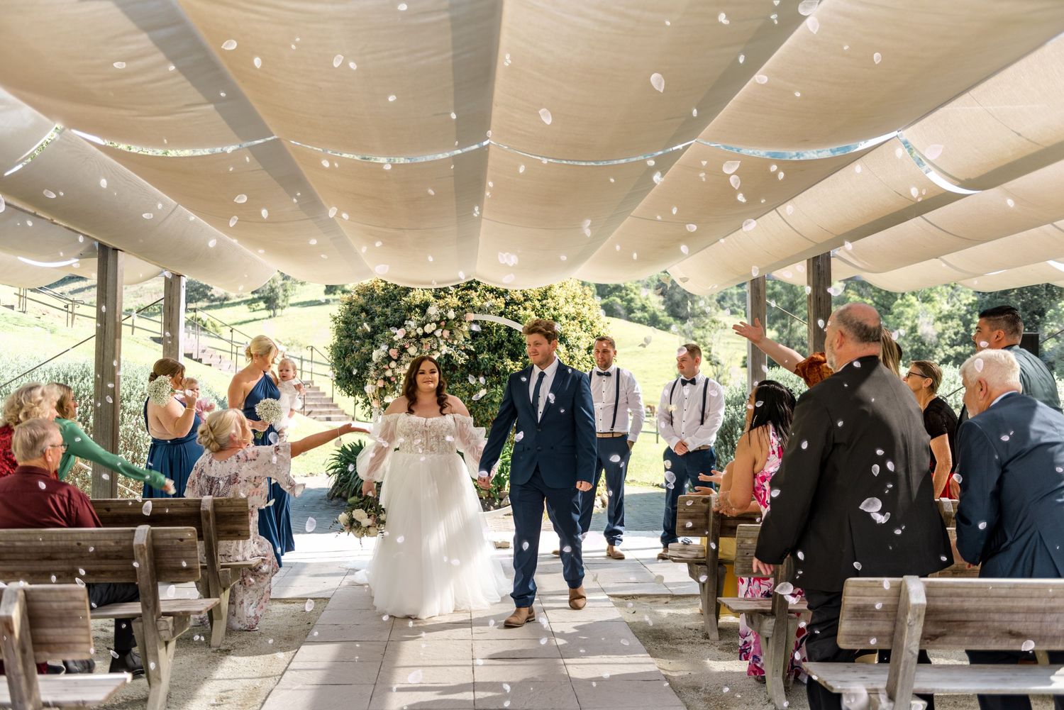 Newlyweds walking down aisle as guests celebrate with confetti under draped canopy at outdoor wedding.