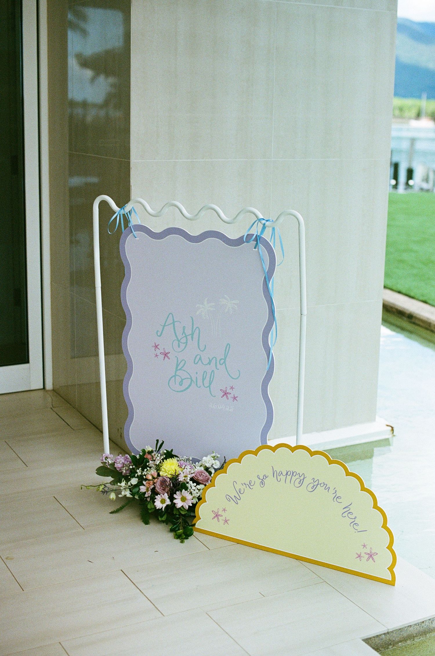 Wedding welcome sign reading Ash and Bill in purple and yellow scalloped frames with floral arrangement.