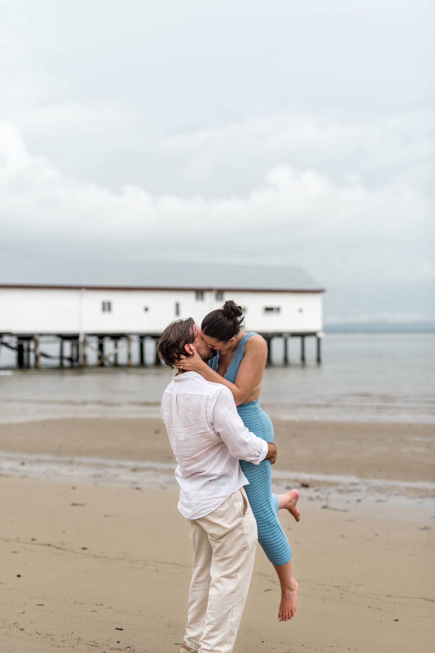 Man lifting woman in blue outfit for a kiss on a sandy beach with white building on pier.
