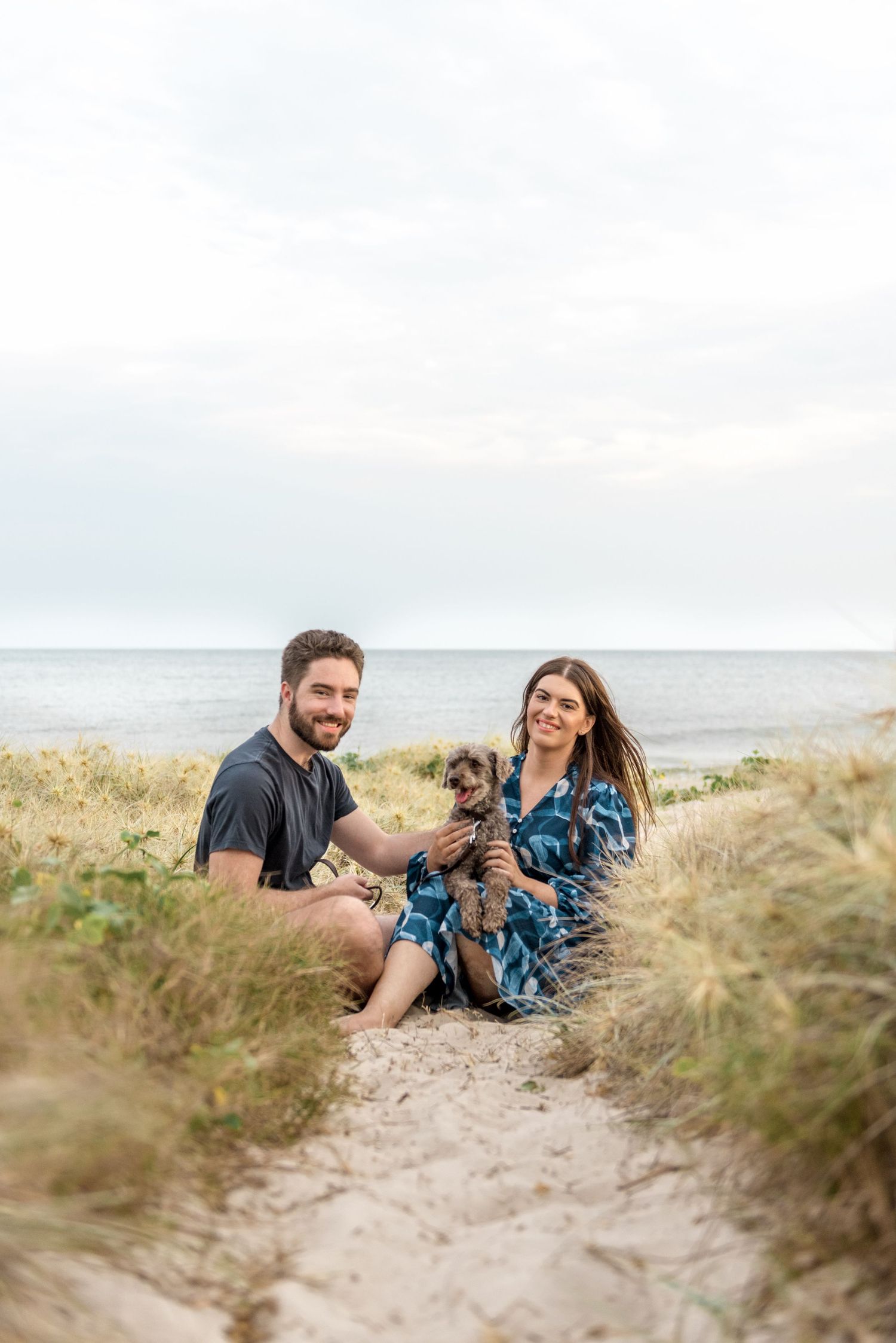 Happy couple sitting on sandy beach path holding small grey dog with ocean view.