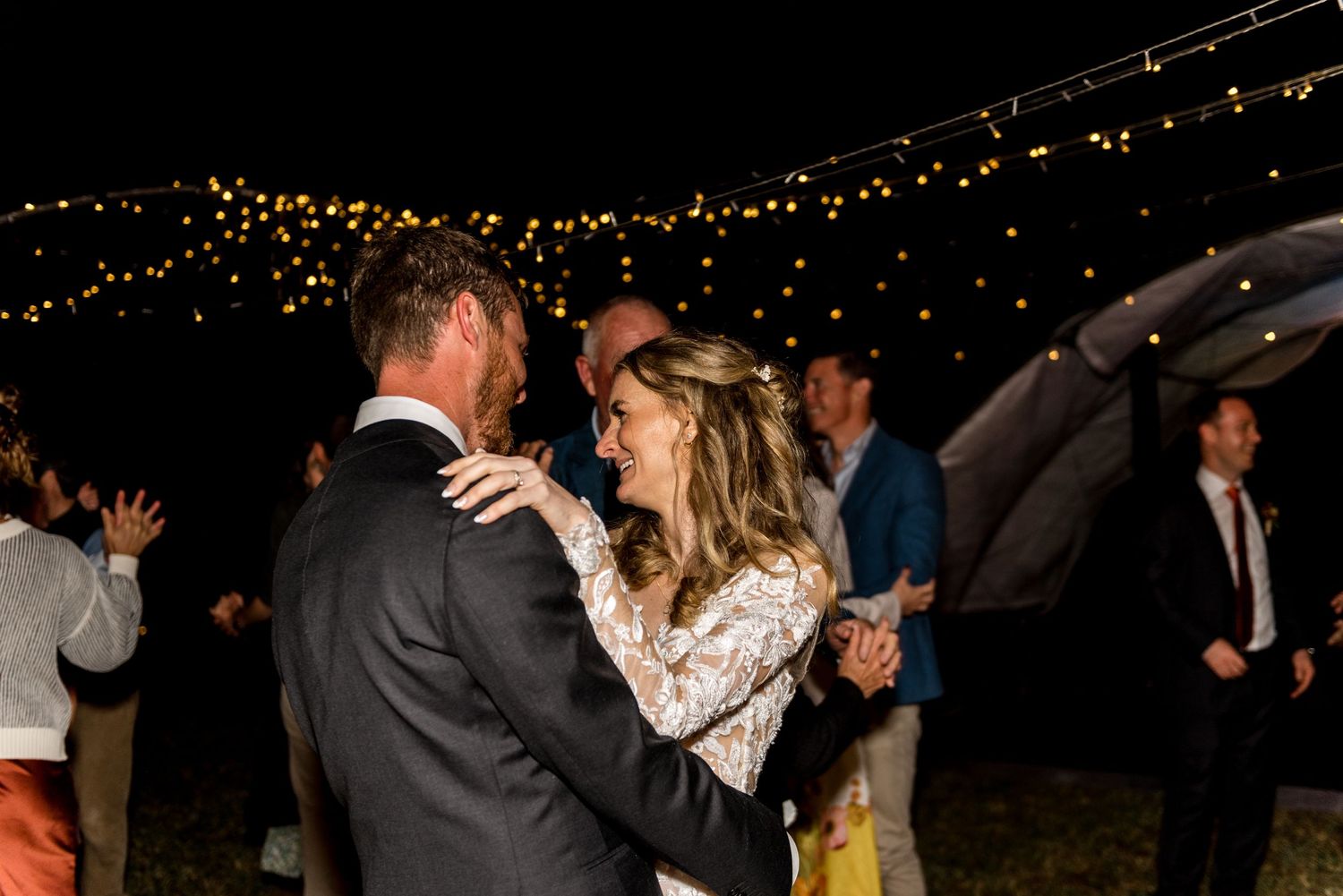 Bride and groom share a joyful dance under twinkling string lights at their outdoor evening wedding reception.