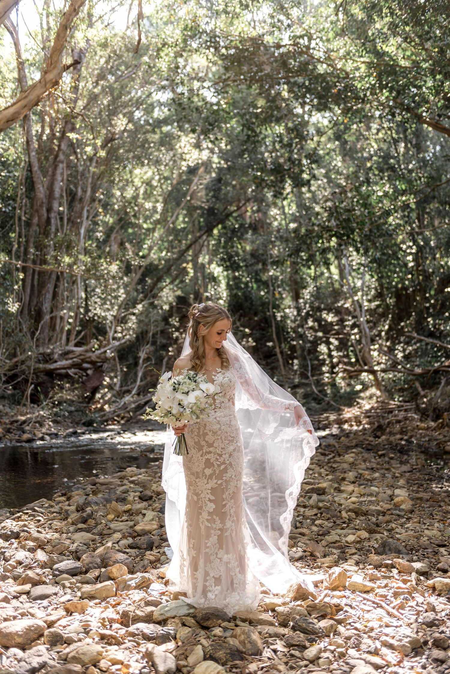 Bride in lace wedding dress holding white bouquet standing on rocky creek bed surrounded by lush forest.