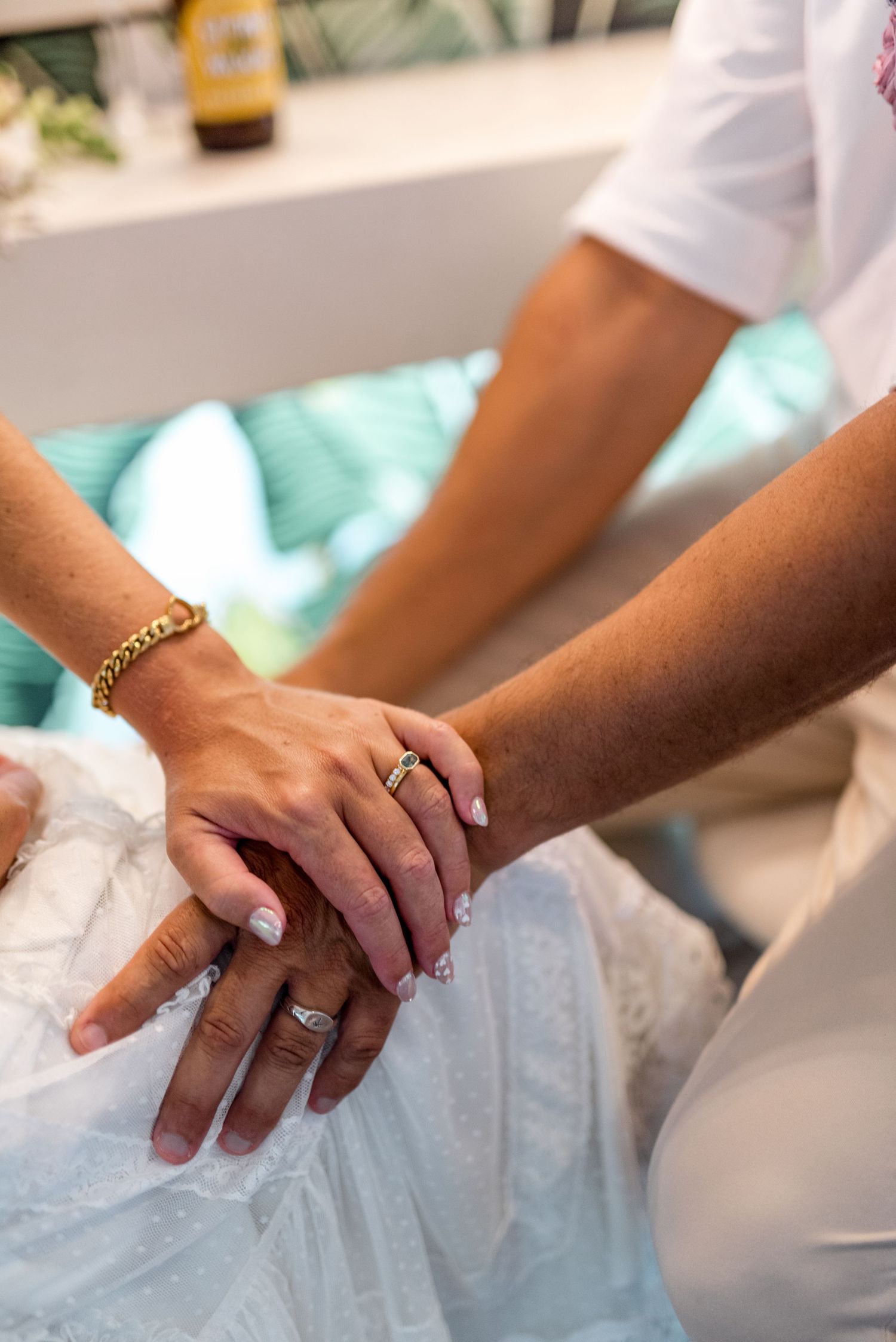Close-up of bride and groom holding hands showing wedding rings, gold bracelet, and white dress.