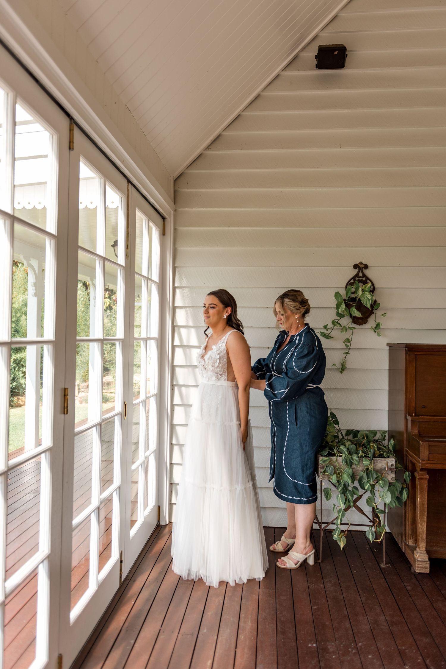 Bride in white lace wedding gown being helped by woman in navy robe on sunlit timber verandah.