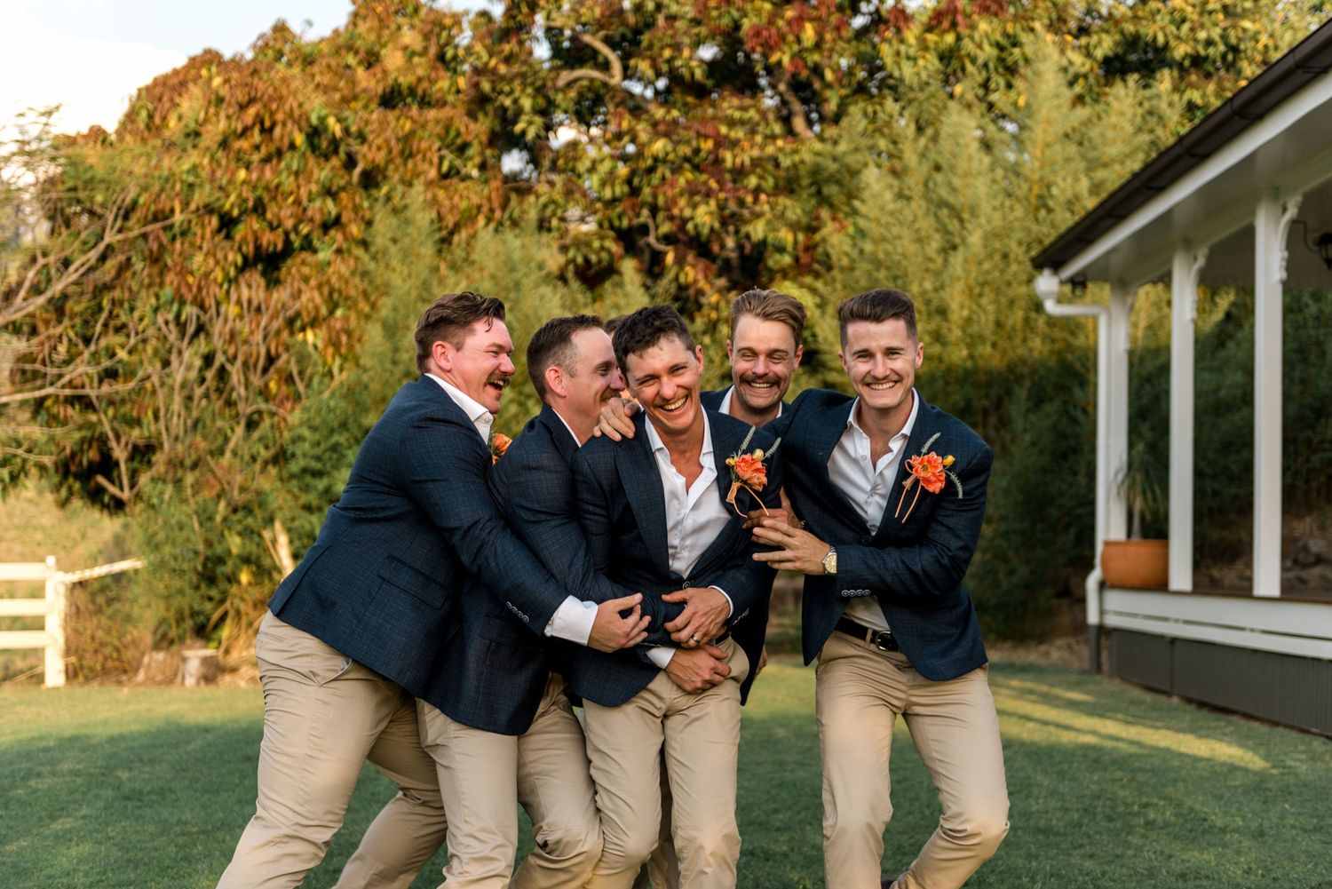 Four groomsmen in navy blazers and khaki pants laughing together on a green lawn.