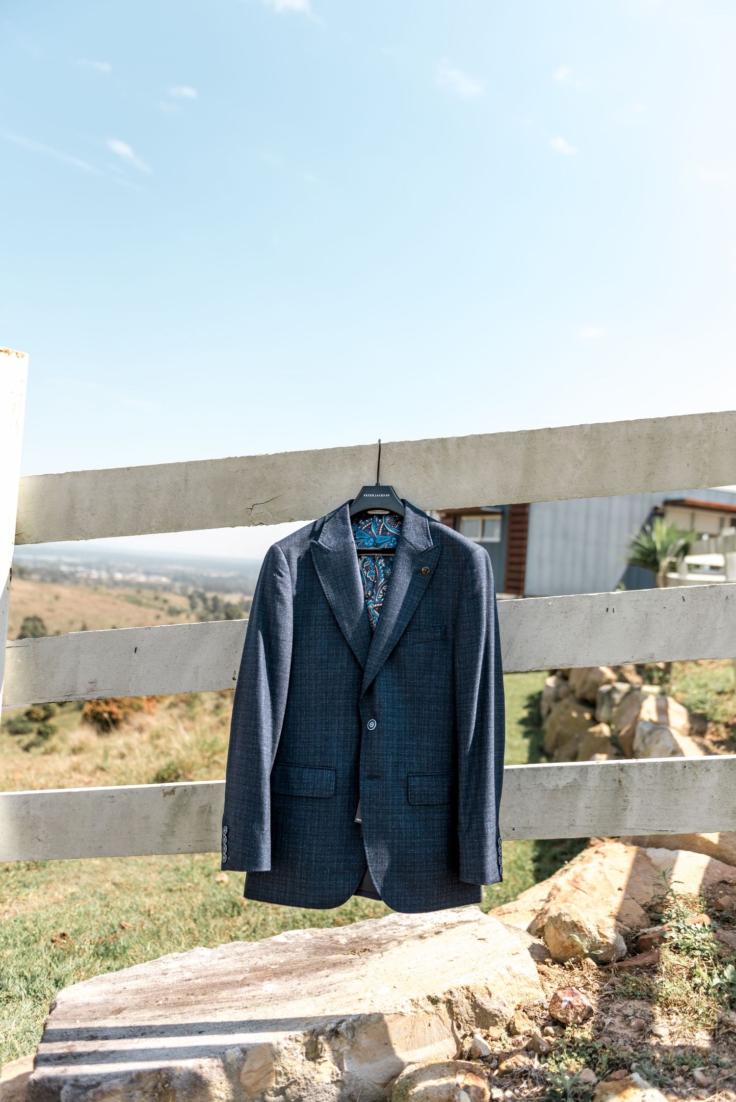 Navy blue checkered groom's blazer hanging on white wooden fence under clear blue sky.