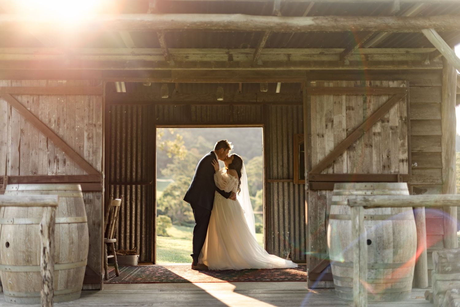 Bride and groom share a romantic kiss silhouetted in the doorway of a rustic wooden barn.