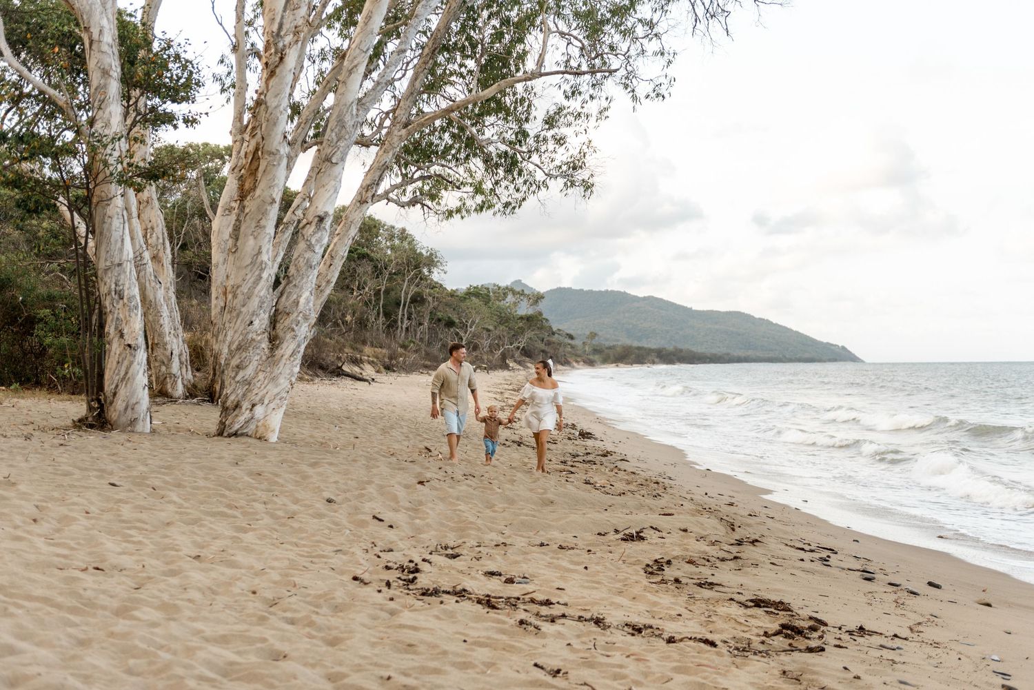A couple walks hand-in-hand with their toddler along a sandy beach with white-trunked trees.