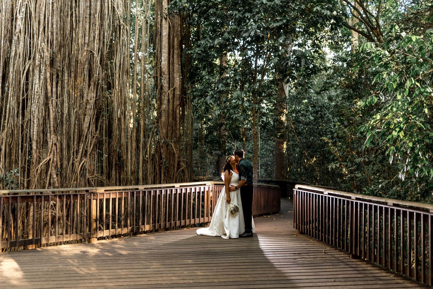 Wedding couple shares a romantic kiss on a wooden bridge surrounded by lush tropical banyan trees.