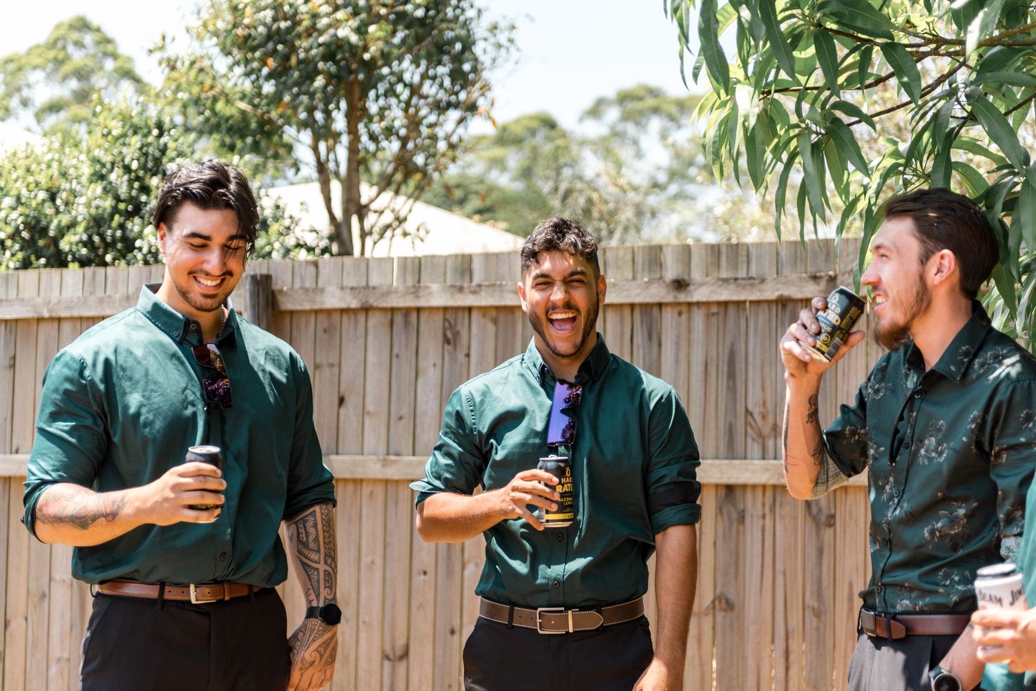 Three groomsmen in matching green shirts laughing and drinking outdoors.