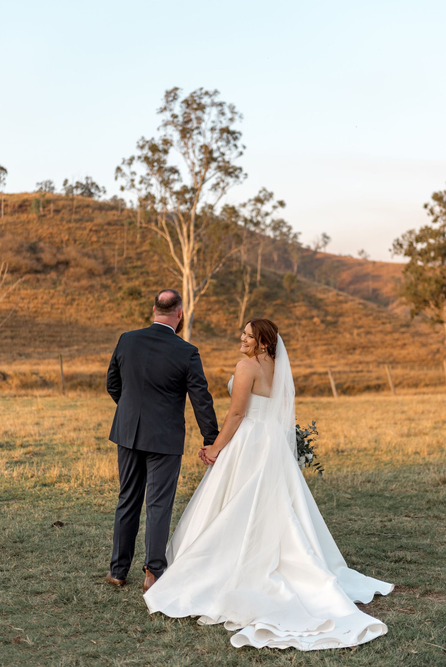 Bride and groom holding hands walking through golden field at sunset with rolling hills behind.