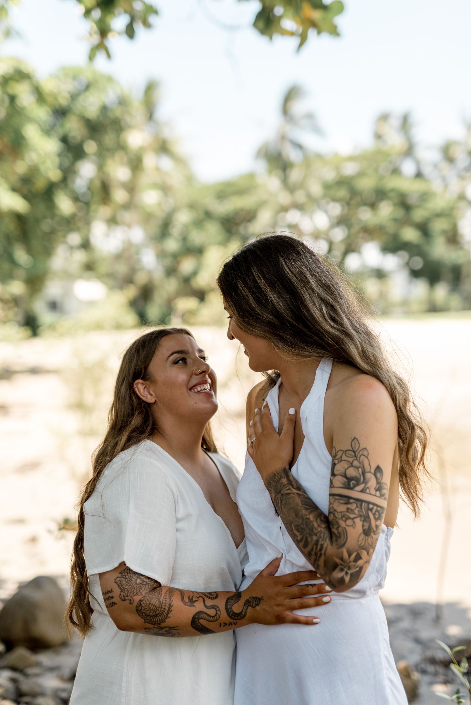 Two tattooed women in white dresses gaze lovingly at each other outdoors with tropical greenery in the background.