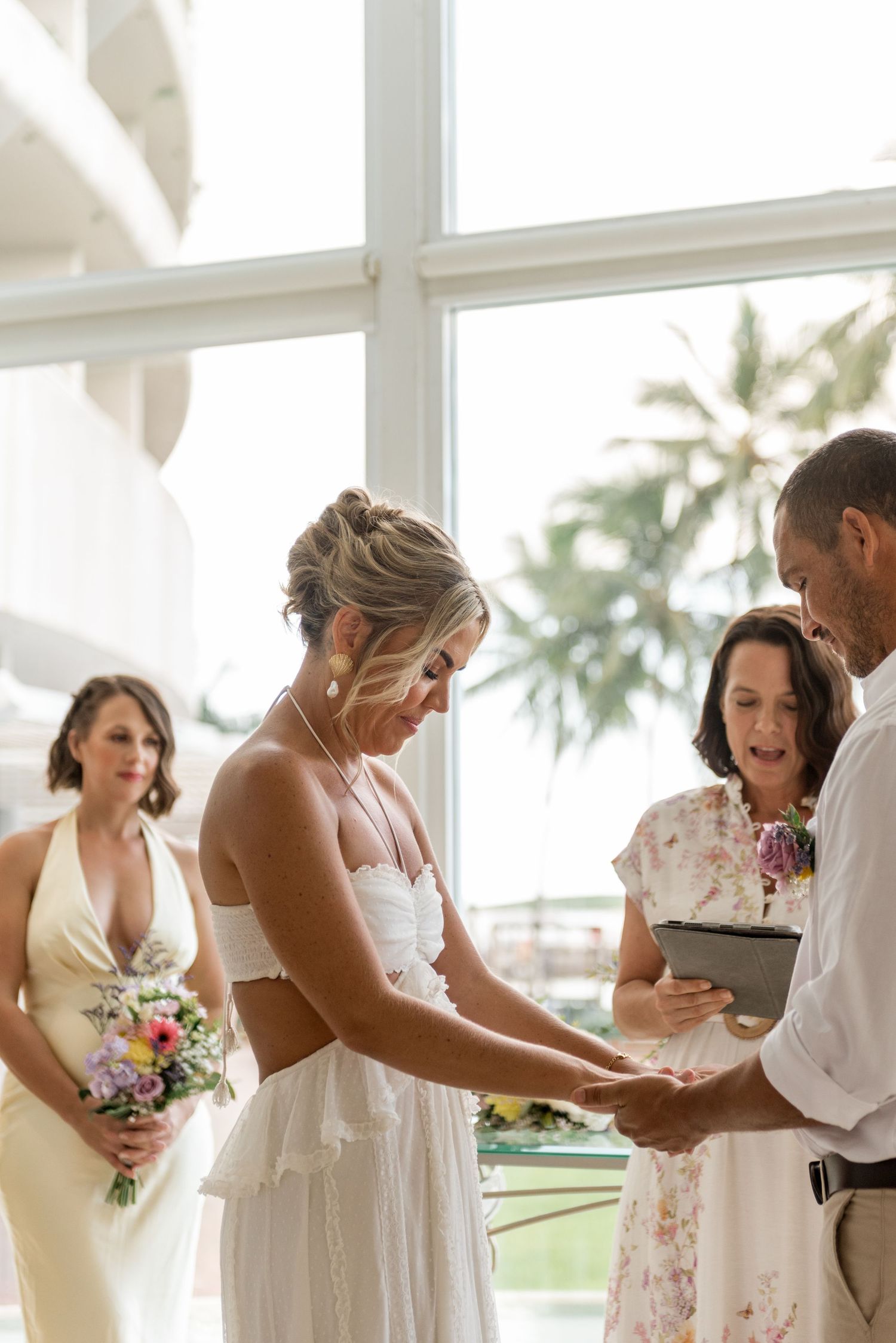 Bride holding bouquet receiving wedding ring during ceremony with bridesmaid and officiant nearby.