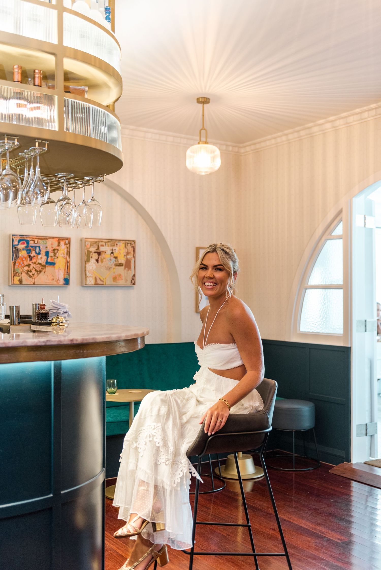 Smiling bride in white wedding dress sits at upscale bar with teal velvet seating and pendant lighting.