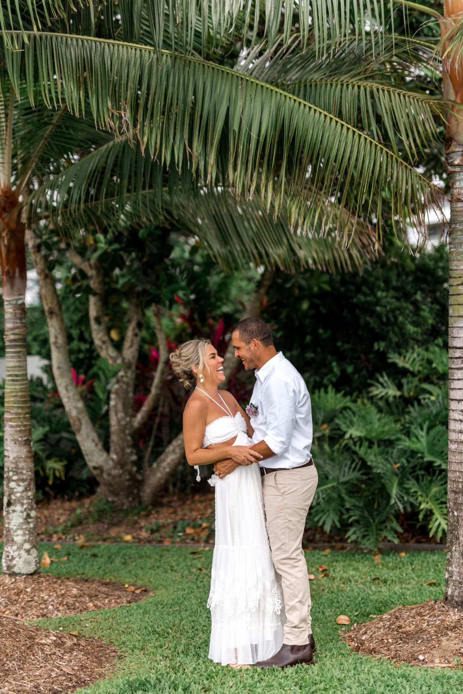 Smiling bride in white dress and groom in white shirt sharing a joyful moment under a palm tree.