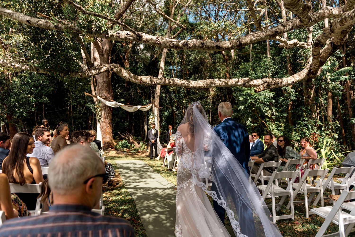 Bride walking down the aisle in lace gown and veil at an outdoor forest wedding ceremony.