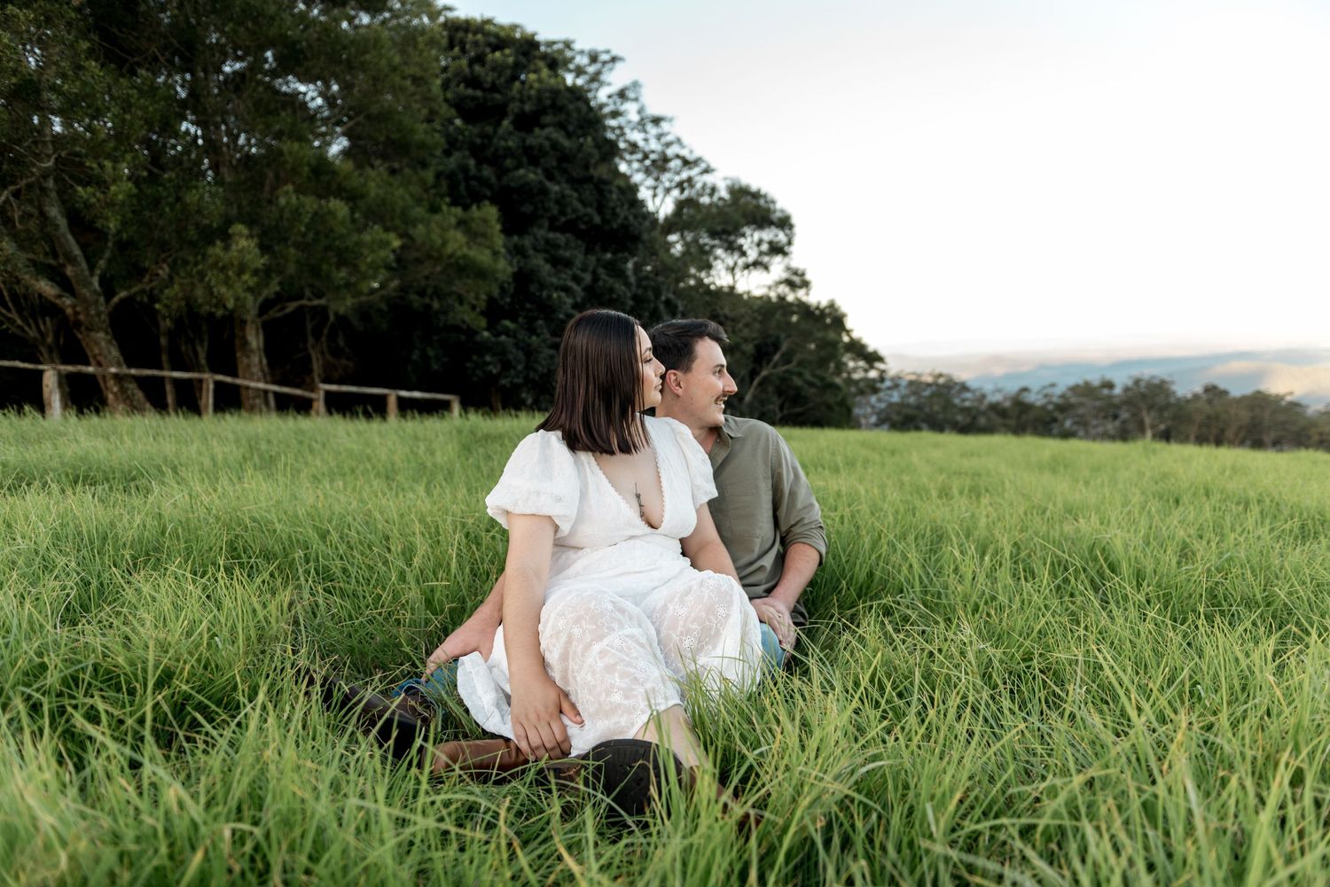 Couple sitting together in lush green field at sunset, woman in white dress leaning against mans shoulder.