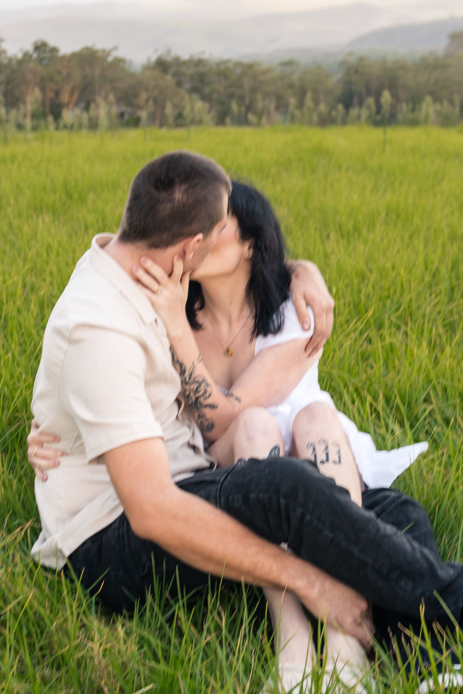 Couple kissing passionately while seated in vibrant green grass field at golden hour.