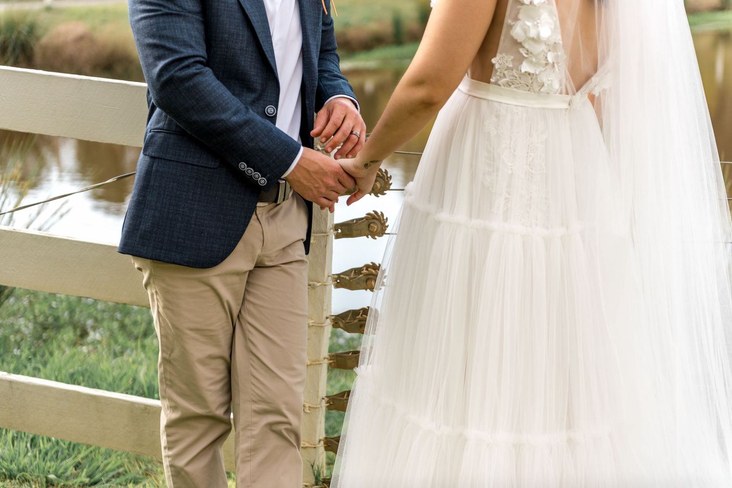 Bride in layered white tulle gown holds grooms hand near a white fence at an outdoor wedding venue.