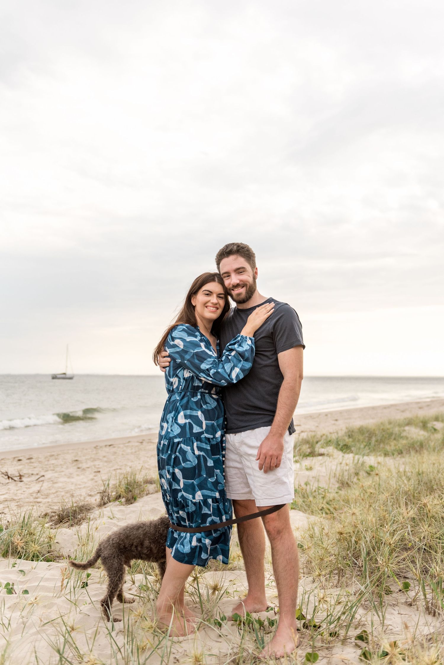 Happy couple embracing on sandy beach with grey dog and ocean backdrop.
