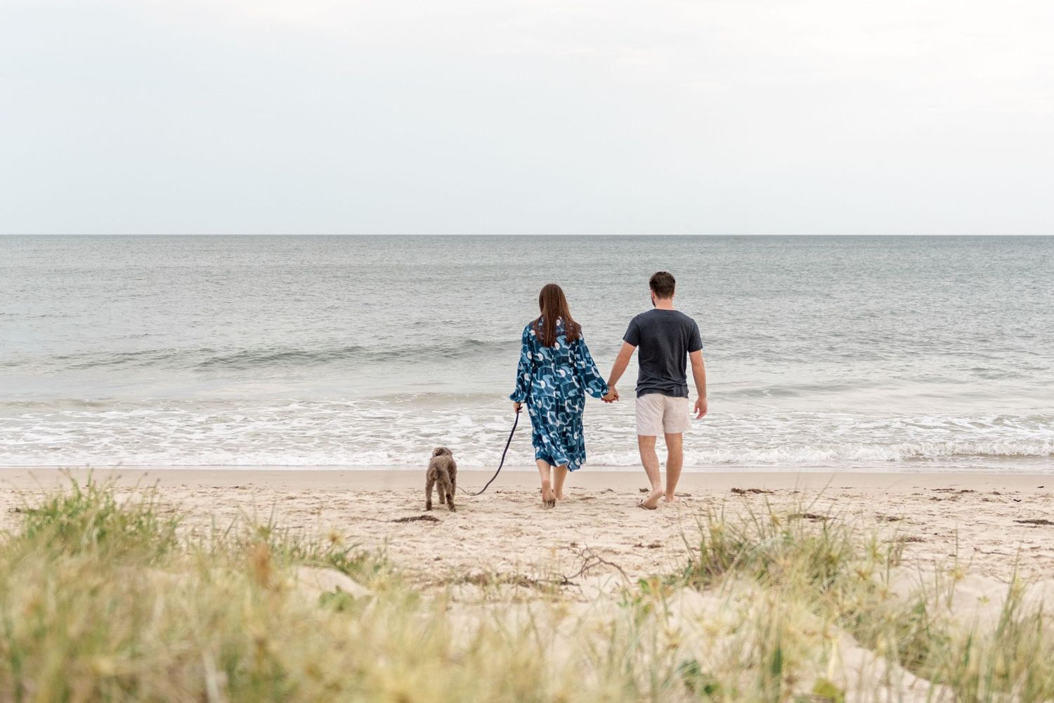 Couple walking hand in hand with their dog along a sandy beach toward the ocean horizon.