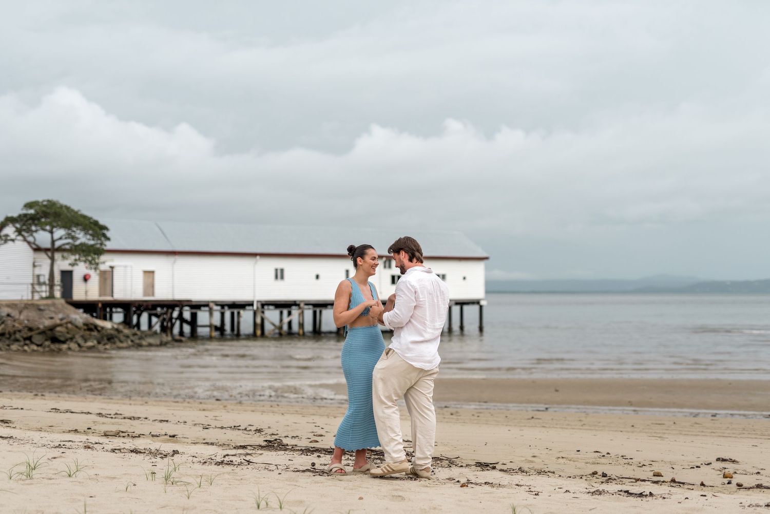 Romantic couple standing close together on a beach with a white pier building visible behind them on an overcast day.