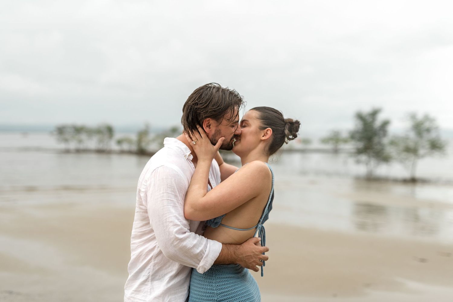 Couple sharing a romantic kiss on a tropical beach under an overcast sky.