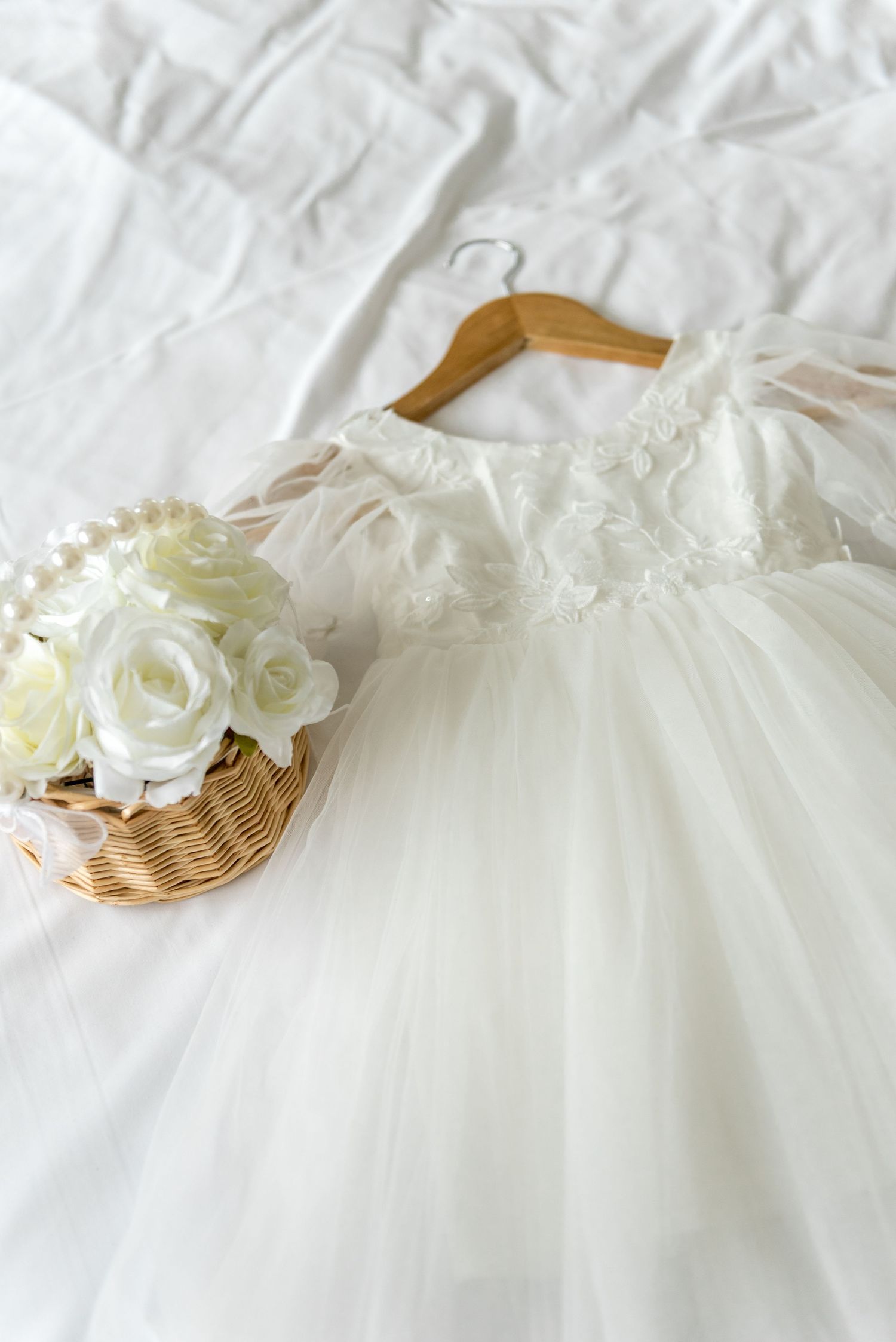 White tulle flower girl dress on a wooden hanger beside a wicker basket of white roses and pearl necklace.