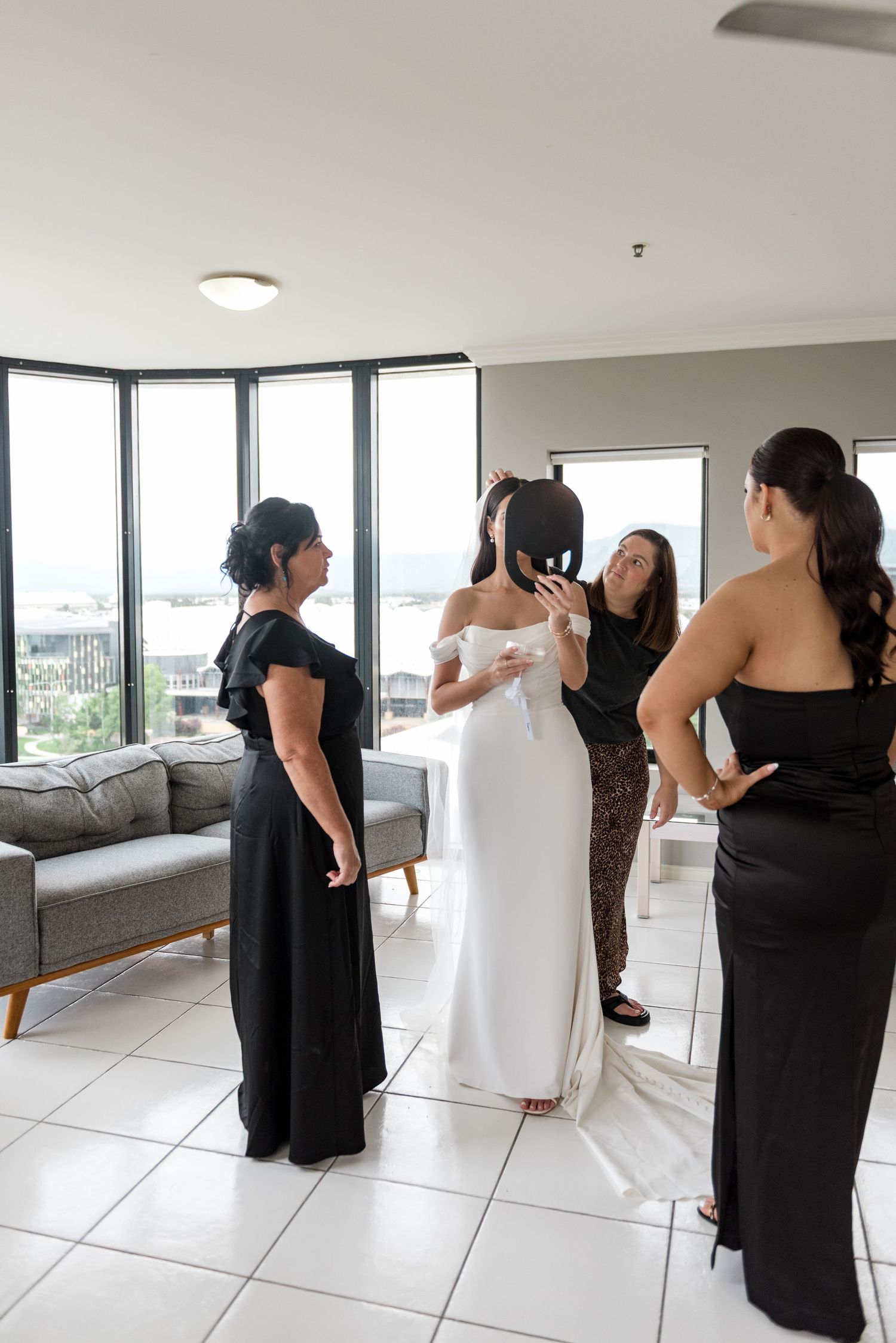 Bridesmaid adjusts bride's white gown as wedding party prepares in a bright modern apartment room.