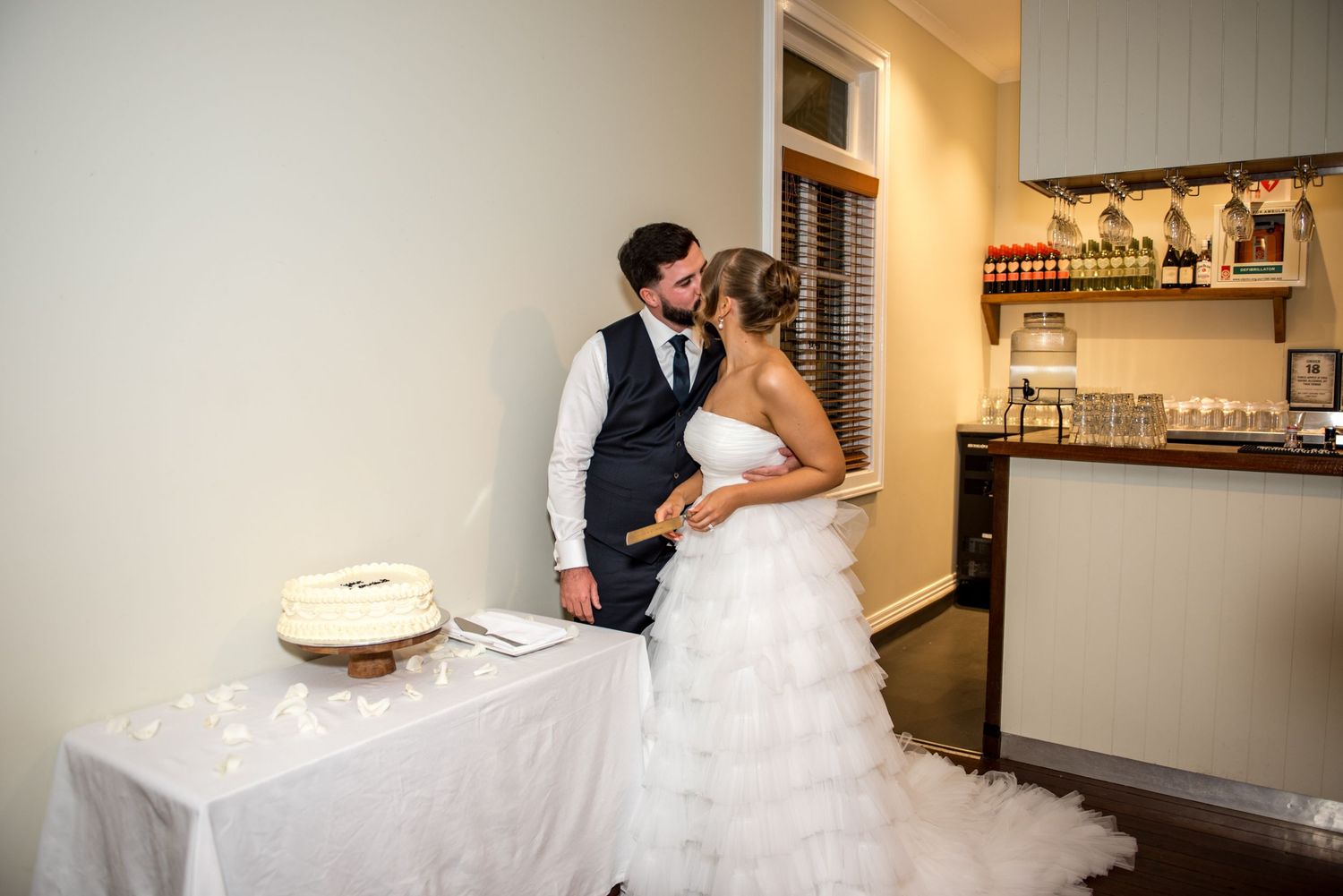 Bride and groom sharing a kiss beside their white wedding cake at the reception.