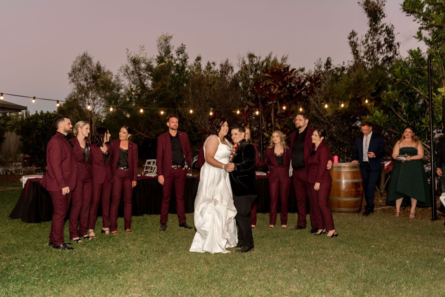 Wedding party in burgundy suits flanking couple during first dance at outdoor evening reception.