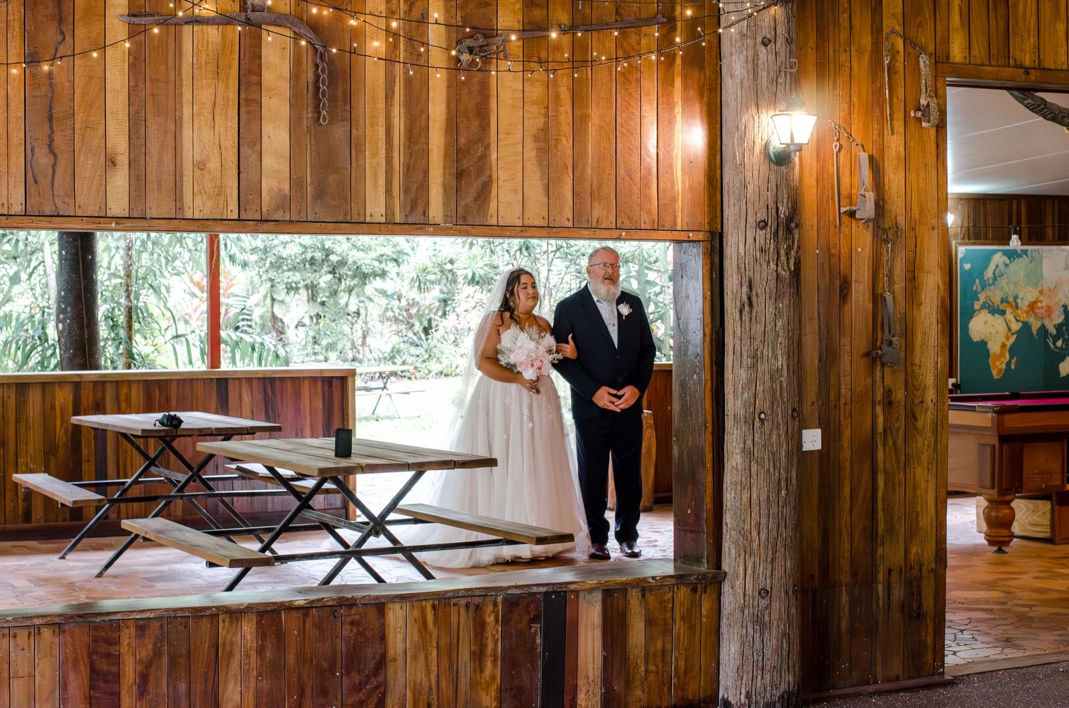Bride in white gown walking with escort through a rustic wooden wedding venue interior.