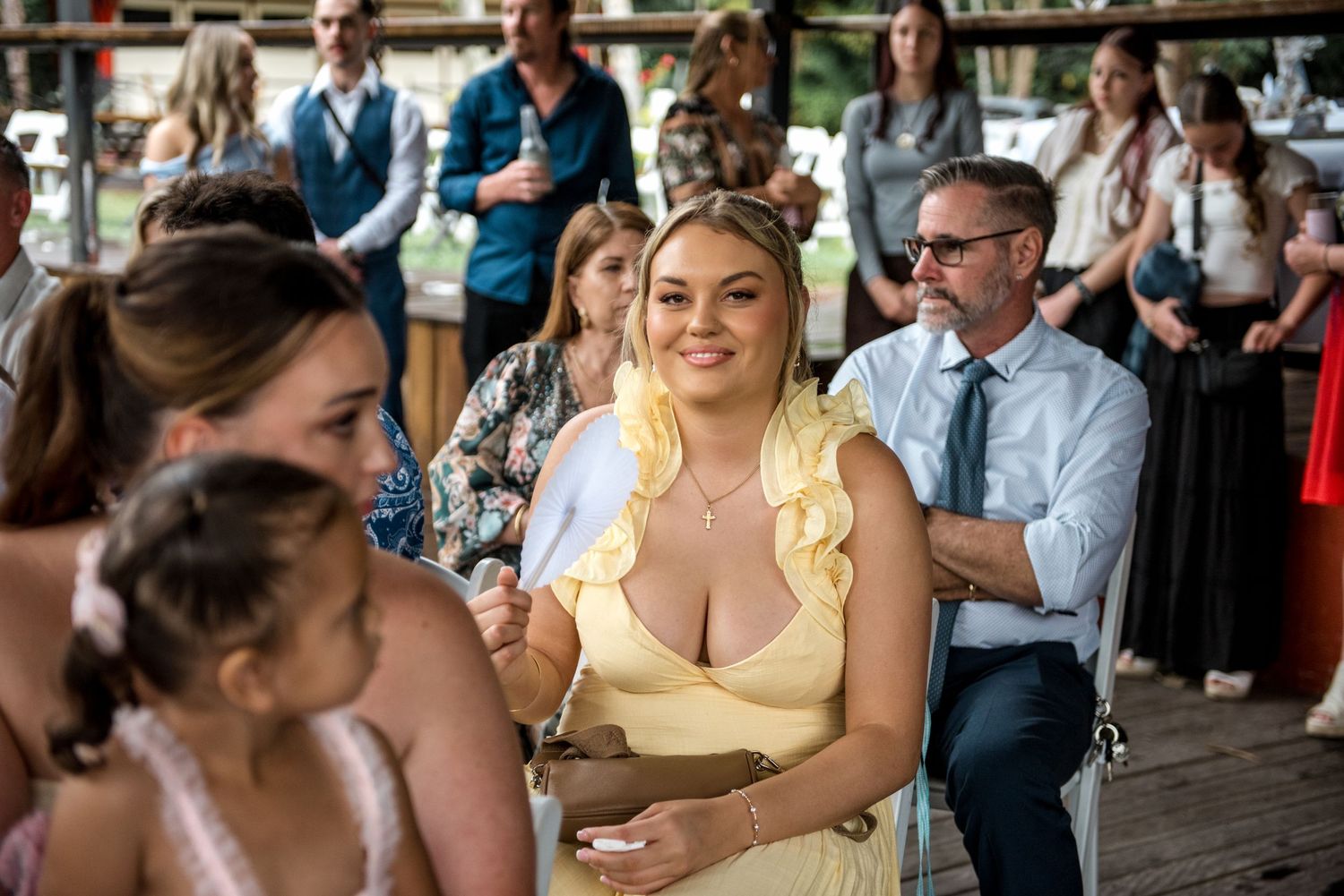Smiling wedding guest in yellow ruffled dress seated at an outdoor wedding ceremony.